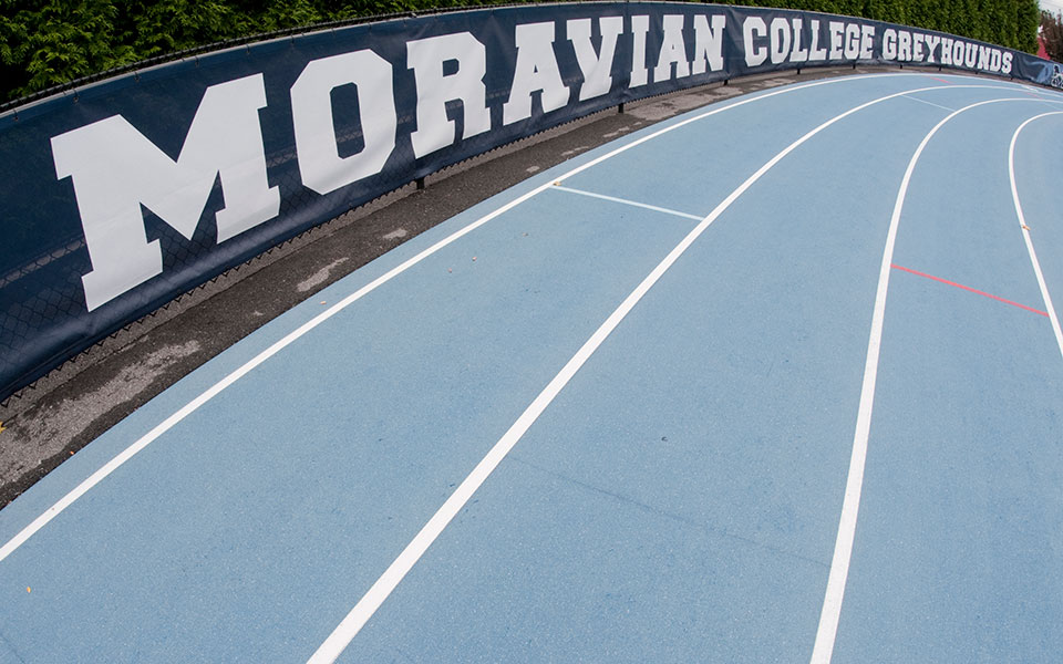 Timothy breidegam track on the first turn with Moravian College Greyhounds banner.